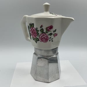 May include: A stovetop espresso maker featuring a cream-coloured ceramic upper chamber decorated with pink rose blossoms and green leaves. The base is constructed from faceted silver-toned metal, and the lid includes a matching ceramic knob handle.