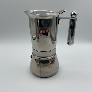 May include: A stainless steel stovetop espresso maker with a handle and a small knob. The maker has a red and white "vev VIGANO" label. The coffee maker is designed to brew espresso on a stovetop.