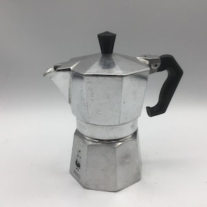 May include: A silver stovetop espresso maker with a black handle and top. The coffee pot has a hexagonal base and a spout for pouring. The brand name is visible on the front of the pot.