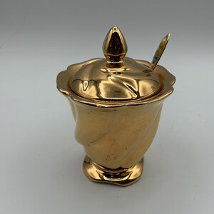 Peut inclure: Un sucrier en céramique doré avec un couvercle et une petite cuillère. Le bol présente un design torsadé unique et un bouton décoratif sur le couvercle. La cuillère a un motif floral.