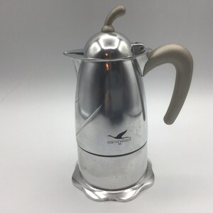 May include: A stainless steel coffee maker with a curved light brown handle and lid handle. The stovetop espresso maker has a logo of a flying bird and text on the front. The base has a scalloped edge.