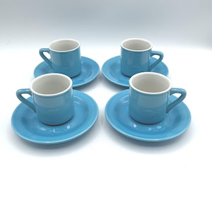 May include: Four matching light blue ceramic espresso cups and saucers. Each cup has a white interior and a curved handle. The saucers are round and the same shade of blue. The set is arranged on a white surface.