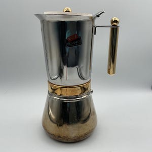 May include: A stainless steel stovetop espresso maker with gold accents. The coffee pot has a gold handle and a gold band around the middle. The brand name "Vev Vigano" is visible on a sticker on the side.