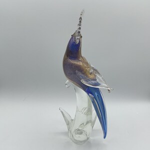 K&ouml;nnte beinhalten: Eine Glasskulptur eines Vogels in Blau, Gold und Klar. Der Vogel hat einen langen, nach oben gerichteten Kamm und einen langen, blauen Schwanz. Die Skulptur ist auf einem klaren Glassockel montiert.