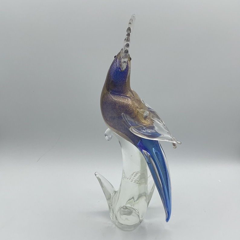 K&ouml;nnte beinhalten: Eine Glasskulptur eines Vogels in Blau, Gold und Klar. Der Vogel hat einen langen, nach oben gerichteten Kamm und einen langen, blauen Schwanz. Die Skulptur ist auf einem klaren Glassockel montiert.