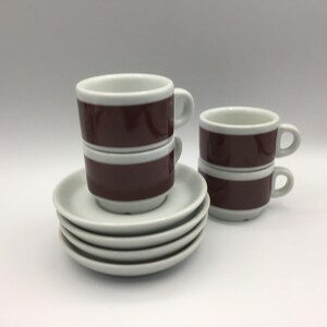 May include: A collection of espresso cups and saucers. The cups are burgundy with white bands and handles, and the saucers are white. The cups are stacked, and the saucers are in a neat pile.