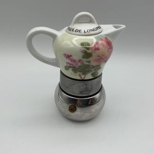 May include: A stovetop espresso maker featuring a white porcelain upper chamber decorated with pink rose illustrations and the text PAUL DE LONGPRE on the lid. The lower base is constructed from polished silver-toned metal with a small brass valve.