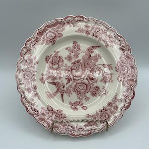 May include: A white ceramic plate with a scalloped edge and a burgundy floral and bird pattern. The plate features a central floral arrangement and a decorative border. The plate is displayed on a metal stand.