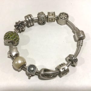 May include: A silver charm bracelet with various decorative beads and charms. The bracelet includes a green beaded charm, a star charm, a pearl, and charms with geometric and floral designs. The bracelet is on a white surface.