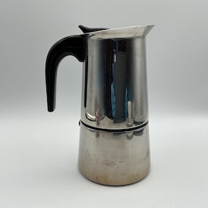 May include: A stainless steel stovetop espresso maker with a black handle. The coffee pot has a classic design with a wide base that tapers upwards. The handle is curved and attached to the side of the pot. The pot is designed to brew coffee on a stovetop.