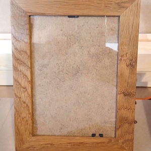 Puede incluir: Un marco de fotos rectangular construido con madera de roble de color marrón claro con un patrón de veta natural visible. El marco presenta esquinas en inglete, un frente de vidrio transparente y un panel trasero de color neutro sujeto por clips.