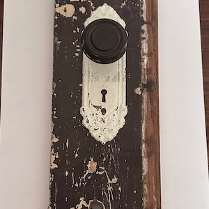 Puede incluir: Una perilla de puerta vintage y una cerradura en una pieza de madera recuperada. La perilla es negra con una placa de porcelana blanca y detalles ornamentados. La madera es marrón oscuro con un acabado desgastado.