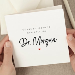 May include: White card with black text that reads "We are so proud to now call you Dr. Morgan" with a small red heart below the text.