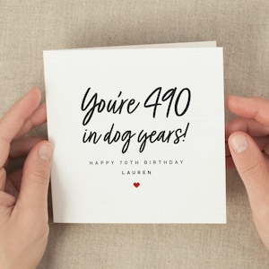 May include: A white square greeting card with black script that reads "You're 490 in dog years!" and "Happy 70th Birthday Lauren" with a small red heart. The card is held by two hands.