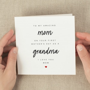 May include: A white Mother's Day card with black script that reads "TO MY AMAZING mom ON YOUR FIRST MOTHER'S DAY AS A grandma I LOVE YOU MOM" with a small red heart. The card is held by two hands.