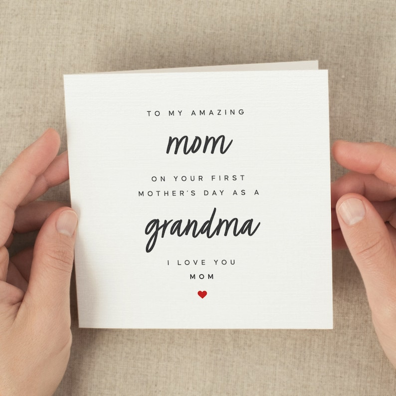 May include: A white Mother's Day card with black script that reads "TO MY AMAZING mom ON YOUR FIRST MOTHER'S DAY AS A grandma I LOVE YOU MOM" with a small red heart. The card is held by two hands.