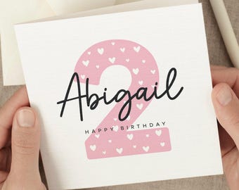 May include: A white birthday card with the name "Abigail" in black script and a pink number "2" filled with white hearts. The words "HAPPY BIRTHDAY" are printed below the number.