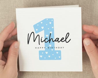 May include: A white birthday card with a blue number one filled with white hearts. The name "Michael" is written in black script, with "HAPPY BIRTHDAY" below. The card is held by two hands.