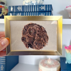 May include: A framed embroidery of a brown dog's head, with a gold frame and a beige background.