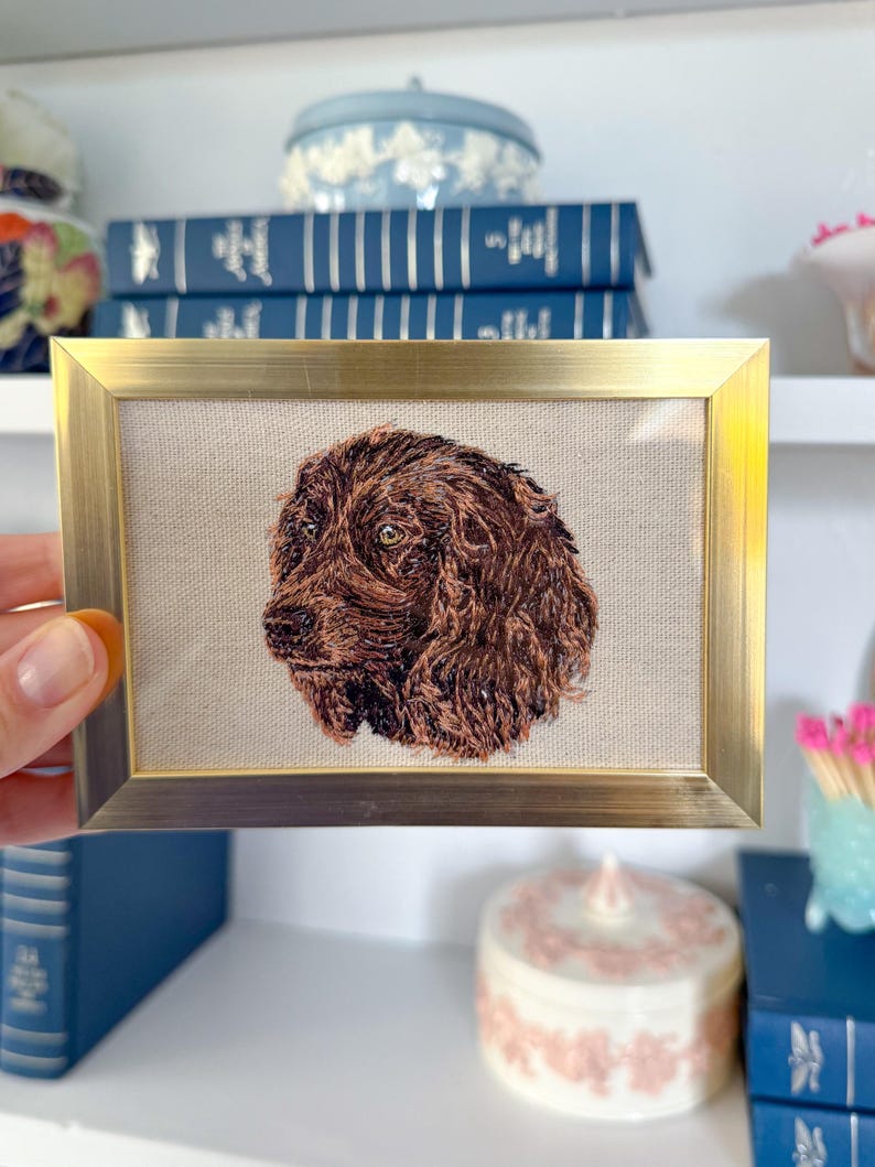 May include: A framed embroidery of a brown dog's head, with a gold frame and a beige background.