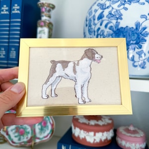 May include: A framed embroidered image of a brown and white Brittany dog on a beige background. The frame is gold.