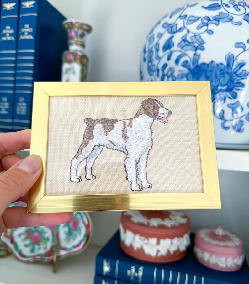 May include: A framed embroidered image of a brown and white Brittany dog on a beige background. The frame is gold.