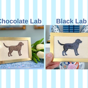 May include: Two framed embroidery pieces depicting Labrador Retrievers. The top piece reads "Black Lab" and shows a black dog. The bottom piece reads "Chocolate Lab" and shows a brown dog.