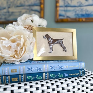 May include: A gold framed embroidery of a brown and white dog on a cream background. The frame is sitting on top of two blue and white patterned books.