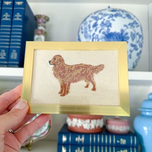 May include: A framed embroidery of a golden retriever dog on a cream-coloured canvas with a gold frame.