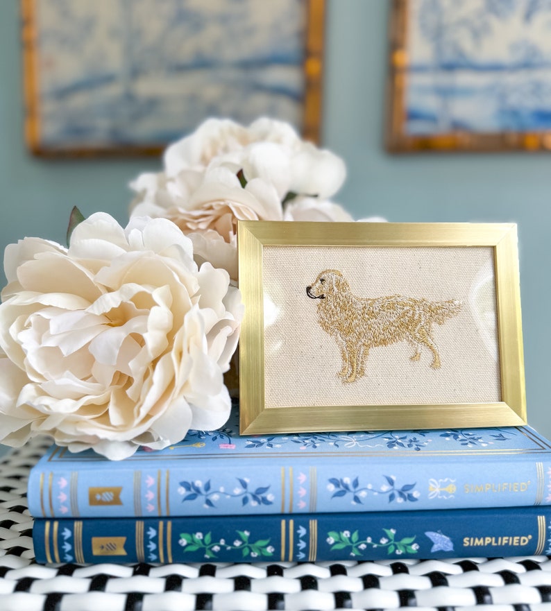 May include: An embroidered picture of a golden retriever dog in a gold frame. The frame is placed on top of two blue and white books with the word "SIMPLIFIED" printed on the spines.