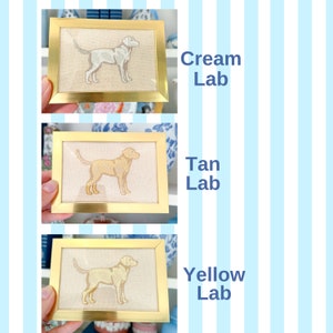 May include: Three framed embroidery pictures of Labrador Retrievers in different colours: cream, tan, and yellow. Each picture has a gold frame and a white background.