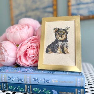 May include: A framed embroidery of a Yorkshire Terrier dog in a sitting position. The embroidery is on a cream-coloured fabric and the frame is gold. The frame is sitting on top of two blue books with the word "SIMPLIFIED" printed on the spines.