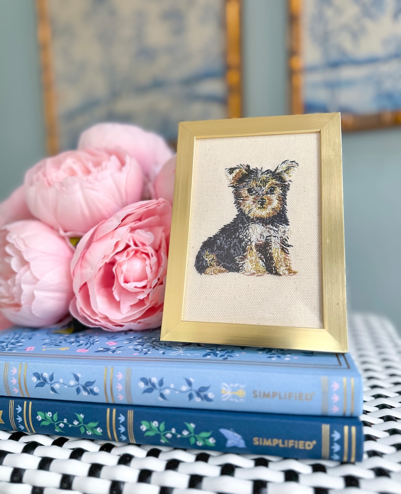 May include: A framed embroidery of a Yorkshire Terrier dog in a sitting position. The embroidery is on a cream-coloured fabric and the frame is gold. The frame is sitting on top of two blue books with the word "SIMPLIFIED" printed on the spines.