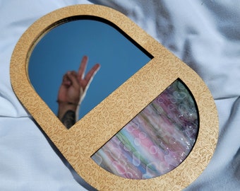 May include: An oval-shaped mirror with a light wood frame featuring a leaf pattern. The mirror reflects a blue sky and a hand making a peace sign. The lower section has a colorful, abstract design.