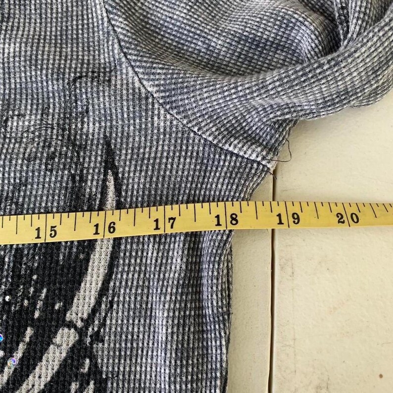 Peut inclure: Un haut &agrave; manches longues en maille gaufr&eacute;e grise avec un imprim&eacute; graphique noir et de petites d&eacute;corations iris&eacute;es. Un m&egrave;tre ruban jaune est plac&eacute; sur la poitrine, indiquant une largeur comprise entre 38 et 51 cm.