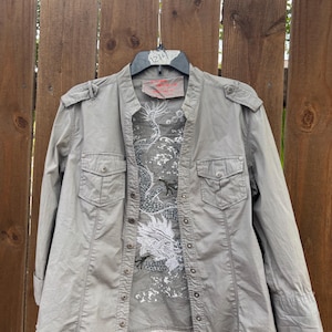 K&ouml;nnte beinhalten: Ein graues Hemd mit Kn&ouml;pfen, Stehkragen und zwei Brusttaschen. Das Hemd hat einen wei&szlig;en und grauen Drachenaufdruck auf der Vorderseite. Das Hemd h&auml;ngt an einem Holzzaun.