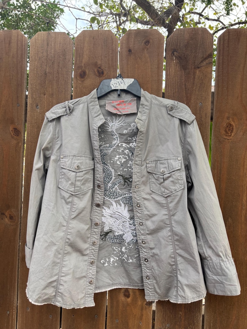 K&ouml;nnte beinhalten: Ein graues Hemd mit Kn&ouml;pfen, Stehkragen und zwei Brusttaschen. Das Hemd hat einen wei&szlig;en und grauen Drachenaufdruck auf der Vorderseite. Das Hemd h&auml;ngt an einem Holzzaun.