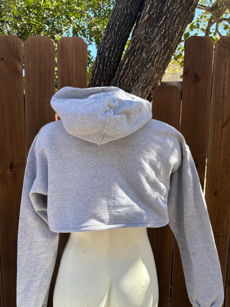 May include: A light gray cropped hoodie with a drawstring hood and long sleeves. The sweatshirt is displayed on a mannequin. The background includes a wooden fence and tree.