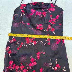 Op de afbeelding: Zwarte satijnen jurk met rode bloemen en cirkelvormige patronen. De jurk heeft dunne bandjes en een getailleerd lijfje. Een gele meetlint ligt over de jurk en toont de afmetingen in centimeters.
