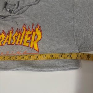 May include: A gray hooded sweatshirt with the word "Thrasher" in yellow and orange flame lettering. The sweatshirt is measured with a yellow measuring tape. The garment has a drawstring hood and long sleeves.