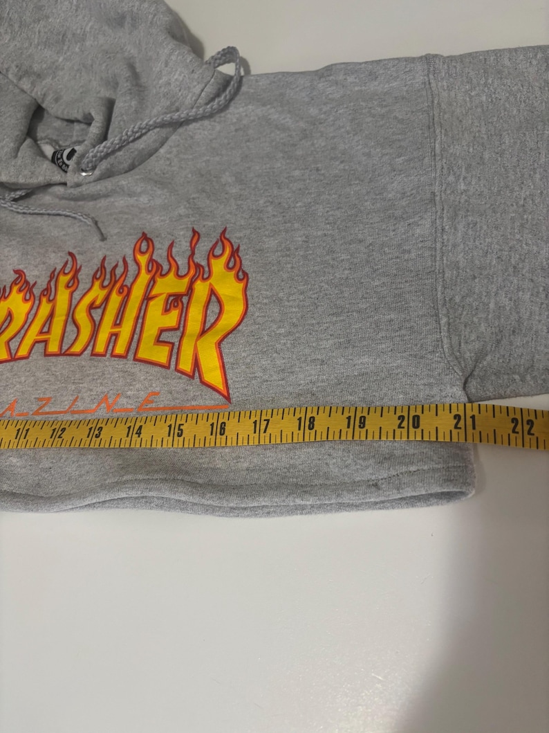 May include: A gray hooded sweatshirt with the word "Thrasher" in yellow and orange flame lettering. The sweatshirt is measured with a yellow measuring tape. The garment has a drawstring hood and long sleeves.
