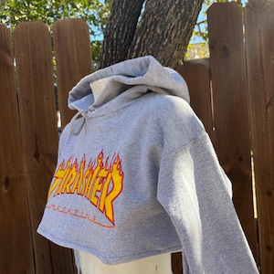 May include: A cropped gray hoodie with the word "Thrasher" in yellow and orange flame lettering. The hoodie has long sleeves and a drawstring hood. The word "Magazine" is printed below the main text.