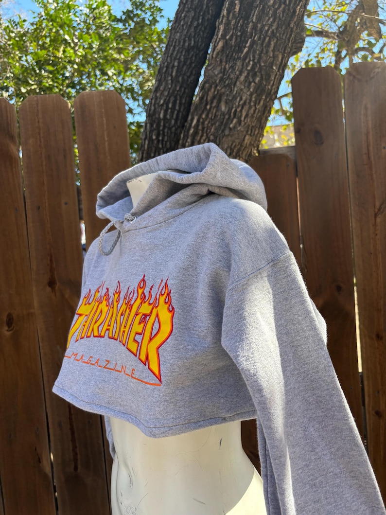May include: A cropped gray hoodie with the word "Thrasher" in yellow and orange flame lettering. The hoodie has long sleeves and a drawstring hood. The word "Magazine" is printed below the main text.