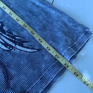 Peut inclure: Un t-shirt bleu et blanc en maille gaufr&eacute;e avec un imprim&eacute; sombre. Un m&egrave;tre ruban jaune est plac&eacute; en diagonale sur le tissu, indiquant une mesure de longueur s'&eacute;tendant de 50,8 cm &agrave; 76,2 cm le long du c&ocirc;t&eacute; du v&ecirc;tement.