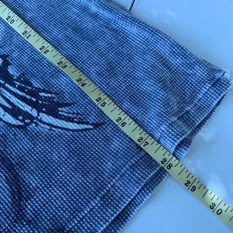 Peut inclure: Un t-shirt bleu et blanc en maille gaufr&eacute;e avec un imprim&eacute; sombre. Un m&egrave;tre ruban jaune est plac&eacute; en diagonale sur le tissu, indiquant une mesure de longueur s'&eacute;tendant de 50,8 cm &agrave; 76,2 cm le long du c&ocirc;t&eacute; du v&ecirc;tement.