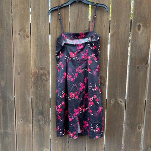 Op de afbeelding: Zwarte jurk met een bloemenpatroon in roze en rode tinten. De jurk heeft dunne bandjes en een vierkante halslijn. Hij hangt aan een zwarte hanger tegen een houten hek.
