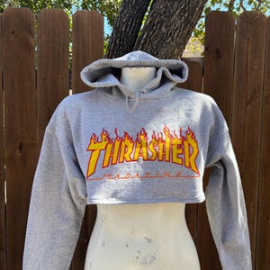 May include: Cropped gray hoodie with the word "Thrasher" in a flame design across the chest. The hoodie has a drawstring hood and long sleeves. The image is of a mannequin wearing the hoodie.