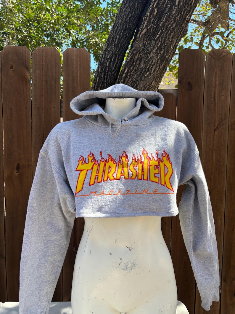 May include: Cropped gray hoodie with the word "Thrasher" in a flame design across the chest. The hoodie has a drawstring hood and long sleeves. The image is of a mannequin wearing the hoodie.