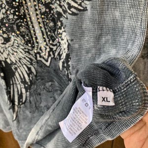Peut inclure: Un t-shirt thermique gris &agrave; manches longues avec un imprim&eacute; graphique noir repr&eacute;sentant des ailes et une croix orn&eacute;e de strass transparents. Le v&ecirc;tement comporte une &eacute;tiquette d'entretien blanche et une &eacute;tiquette de taille XL &agrave; l'int&eacute;rieur.