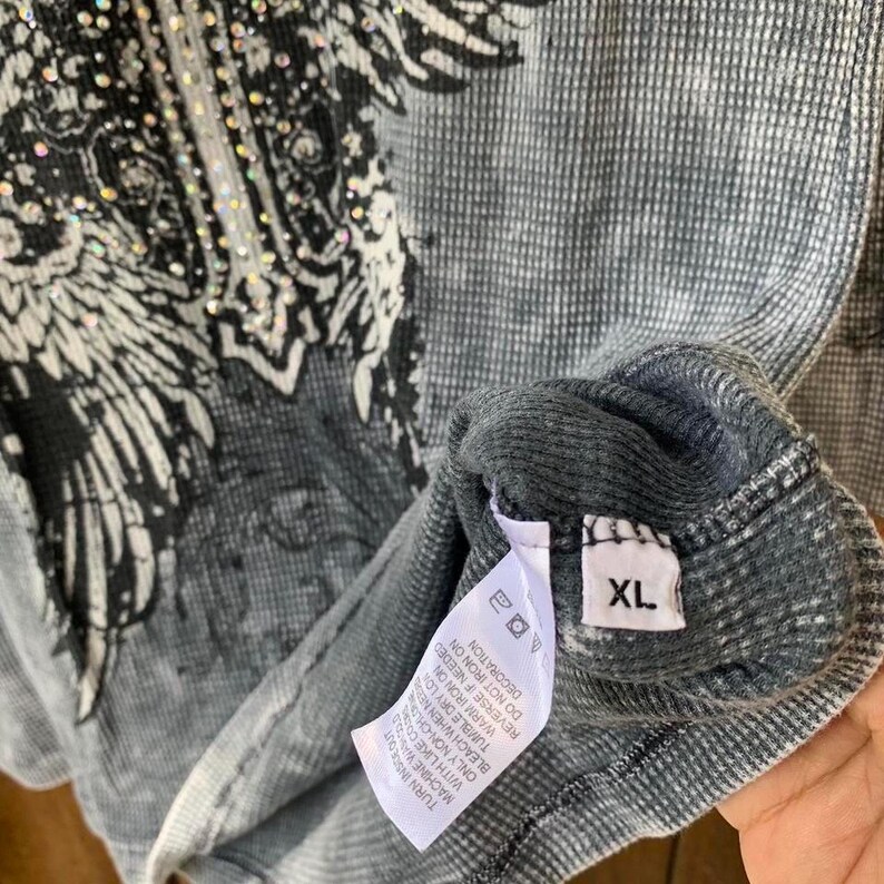 Peut inclure: Un t-shirt thermique gris &agrave; manches longues avec un imprim&eacute; graphique noir repr&eacute;sentant des ailes et une croix orn&eacute;e de strass transparents. Le v&ecirc;tement comporte une &eacute;tiquette d'entretien blanche et une &eacute;tiquette de taille XL &agrave; l'int&eacute;rieur.